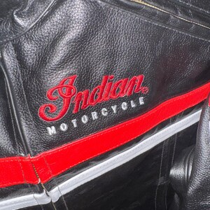 May include: Black leather motorcycle jacket with red and white accents. The jacket features the embroidered text "Indian MOTORCYCLE" in red. A red stripe runs across the chest and a zipper is visible.