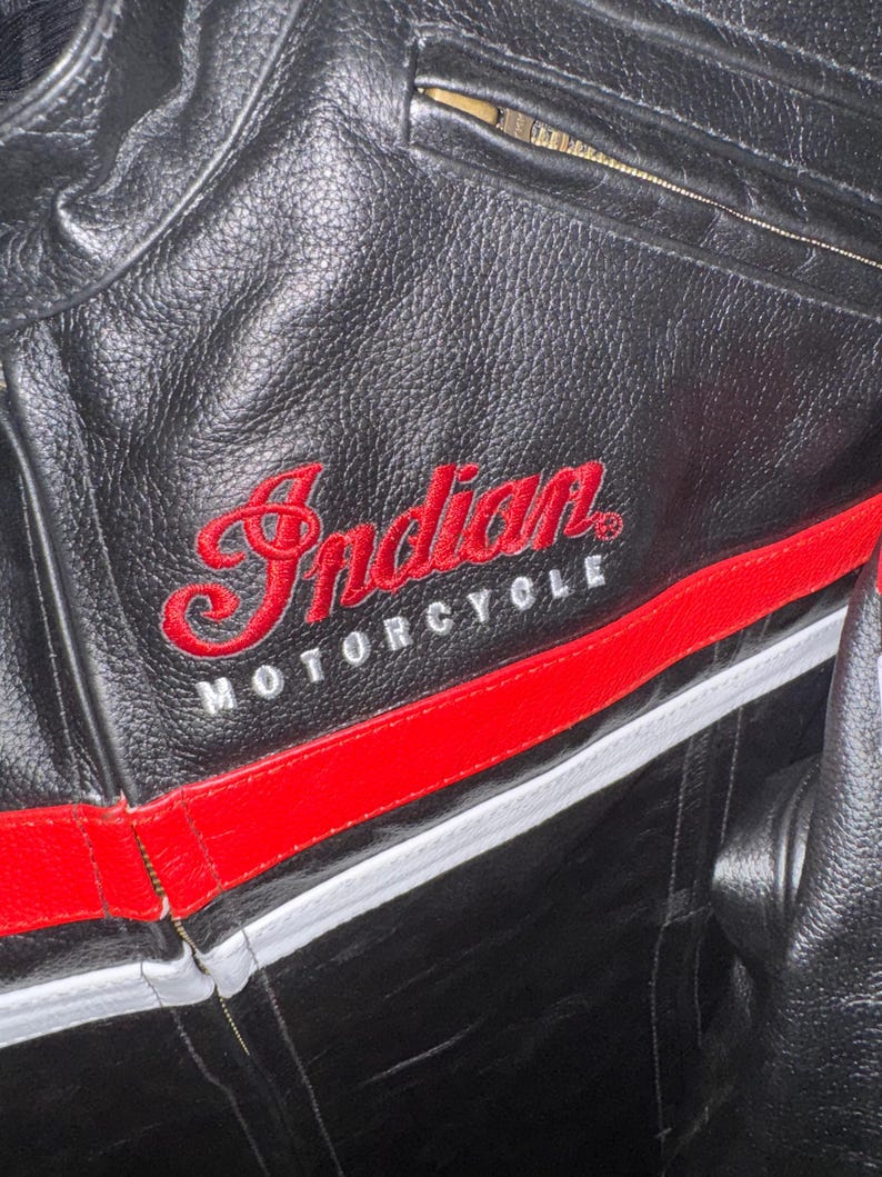 May include: Black leather motorcycle jacket with red and white accents. The jacket features the embroidered text "Indian MOTORCYCLE" in red. A red stripe runs across the chest and a zipper is visible.