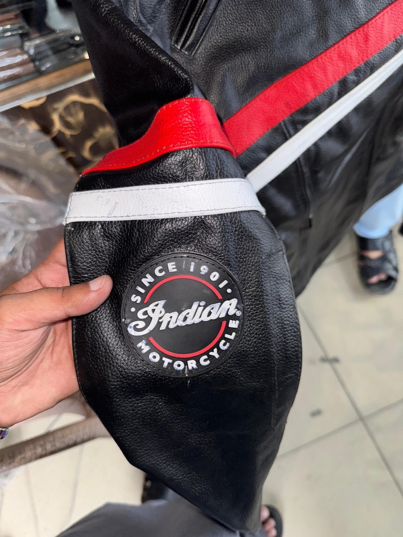 May include: Black leather motorcycle jacket with red and white accents. Features a circular patch with "Indian Motorcycle" text and the words "Since 1901". The jacket has a classic design with a focus on durability and style.