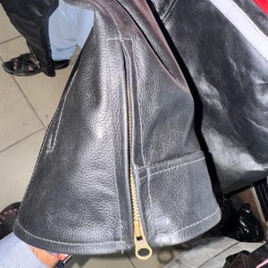 May include: Black leather jacket with a gold zipper and a red and white stripe detail. The jacket has a textured surface and a classic design, suitable for various occasions.