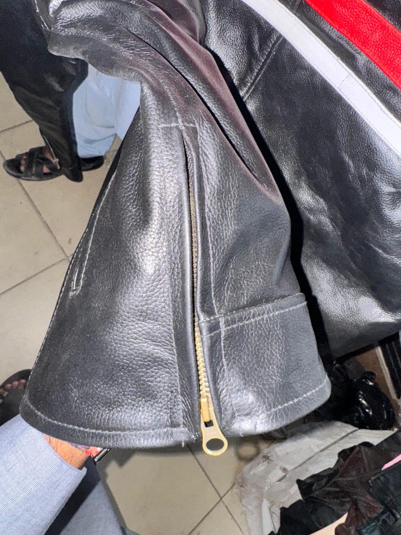 May include: Black leather jacket with a gold zipper and a red and white stripe detail. The jacket has a textured surface and a classic design, suitable for various occasions.