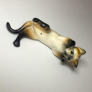 May include: A ceramic figurine of a Siamese cat lying on its back with its paws in the air. The cat is painted in shades of white, brown, and black.