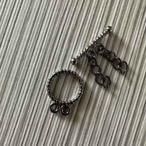 May include: A silver-toned toggle clasp set, featuring a textured ring with two loops and a bar with a twisted design. A small chain hangs from the bar. The clasp is designed for jewelry making.