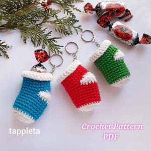 May include: Three crochet keyrings in blue, red, and green, each resembling a small stocking with a white cuff and a white star. The keyrings hold wrapped sweets. The text "tappleta" and "Crochet Pattern PDF" are visible. The keyrings are attached to silver rings.