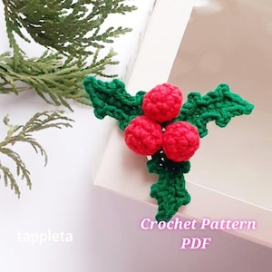 May include: A crocheted holly sprig with three red berries and green leaves. The text "Crochet Pattern PDF" is visible. The sprig is placed on a white surface, with a sprig of green foliage in the background.