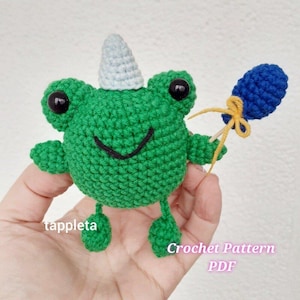 May include: A green crocheted frog with a grey horn, black eyes, and a smiling mouth. It holds a blue crocheted balloon with a yellow string. The text "tappleta" and "Crochet Pattern PDF" are visible.