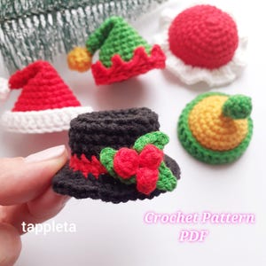 Pode incluir: Uma coleção de chapéus de crochê em miniatura em vários designs festivos. Inclui um gorro de Papai Noel vermelho, um chapéu de elfo verde, um chapéu alto preto com detalhes vermelhos e azevinho e uma bola vermelha com acabamento branco. O texto "Crochet Pattern PDF" também é visível.