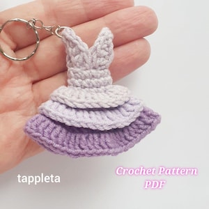 May include: A handmade crochet dress keychain in shades of lavender and purple. The dress features a tiered skirt and a V-neck bodice, attached to a silver keyring. The text "tappleta" and "Crochet Pattern PDF" are visible.