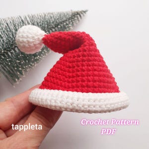 May include: A small, red and white crocheted Santa hat with a white pom-pom. The hat has a white brim and is held in a hand. The text "tappleta" is on the left, and "Crochet Pattern PDF" is on the right.
