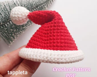 May include: A small, red and white crocheted Santa hat with a white pom-pom. The hat has a white brim and is held in a hand. The text "tappleta" is on the left, and "Crochet Pattern PDF" is on the right.