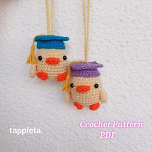 May include: Two handmade crochet duck ornaments, each wearing a graduation cap. The ducks are beige with orange beaks and feet, and black eyes. One cap is blue, the other purple, both with yellow tassels. The text "tappleta" and "Crochet Pattern PDF" are visible.