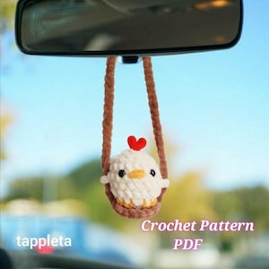 Op de afbeelding: Een gehaakte kippenauto-hanger die aan een achteruitkijkspiegel hangt. De witte kip heeft een rood hart en zit in een bruine schommel. De tekst "Crochet Pattern PDF" en "tappleta" zijn zichtbaar. Een decoratief item voor een voertuig.