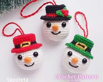 May include: Three crocheted snowman decorations with red, black, and green hats. Each snowman has a carrot nose, black eyes, and a smiling mouth. The decorations have red hanging loops. Text on the image says "tappleta" and "Crochet Pattern PDF".