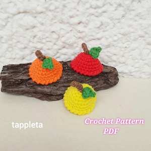 May include: Three crocheted apple ornaments in orange, red, and yellow, each with a brown stem and green leaf. The apples are arranged on a piece of wood. The text "tappleta" and "Crochet Pattern PDF" are visible.
