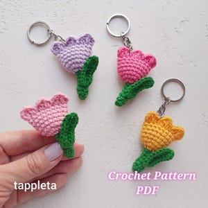 May include: Four crochet flower keychains in shades of pink, purple, and yellow, each with a green stem and attached to a silver keyring. The keychains are handmade and feature a detailed crochet pattern. The text "Crochet Pattern PDF" is visible.
