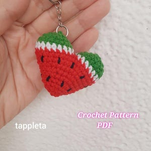 May include: A heart-shaped crochet keychain resembling a watermelon slice. The keychain features red yarn with black seed details, green rind, and white accents. The text "Crochet Pattern PDF" and the seller's name "tappleta" are also visible.