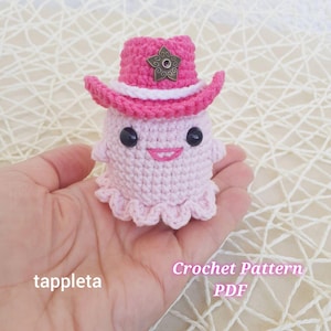 May include: A handmade, light pink crochet ghost wearing a pink cowboy hat with a star-shaped embellishment. The ghost has black eyes and a smiling mouth. The text "tappleta" and "Crochet Pattern PDF" are visible.