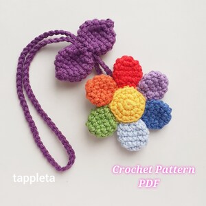 Peut inclure: Une fleur au crochet faite à la main avec une feuille et un cordon violets. La fleur a des pétales de différentes couleurs : rouge, orange, jaune, vert, bleu clair et violet. Le texte "tappleta" et "Crochet Pattern PDF" sont visibles.