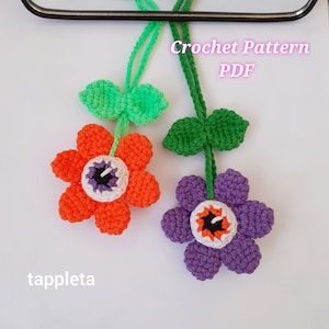 May include: Two crochet flower ornaments with green stems and leaves. One flower is orange, the other is purple. Each flower has a white and black centre, resembling an eye. The text "Crochet Pattern PDF" is visible.