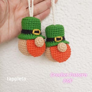 May include: Two handmade crochet leprechaun ornaments. Each ornament features a green hat with a black band and gold buckle, an orange body, and a tan beard. The ornaments are suspended by a white string. The text "tappleta" and "Crochet Pattern PDF" are visible.