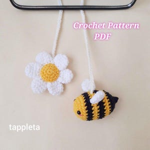 May include: Two crocheted ornaments: a white daisy with a yellow center and a yellow and black bee with white wings. The text "Crochet Pattern PDF" is visible. The word "tappleta" is also visible.