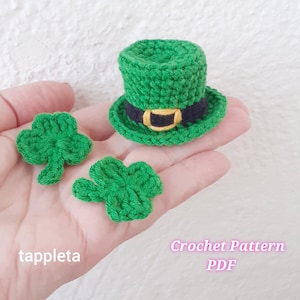 Puede incluir: Primer plano de decoraciones de ganchillo del Día de San Patricio. Un sombrero verde de duende con una banda negra y una hebilla dorada está junto a dos tréboles verdes. El texto "tappleta" y "Crochet Pattern PDF" son visibles.