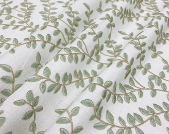 May include: White fabric with an embroidered pattern of green leaves and stems. The leaves have a detailed, textured appearance, and the stems are a muted gold color. The fabric is folded, creating shadows and depth.