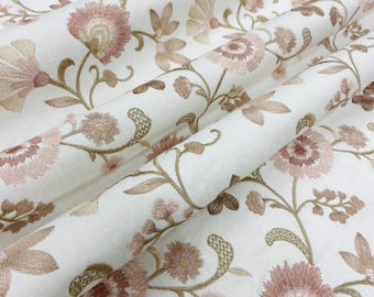 May include: Close-up of a white fabric with an embroidered floral pattern. The design features pink and tan flowers with green stems and leaves. The fabric is folded, showing the texture and detail of the embroidery.