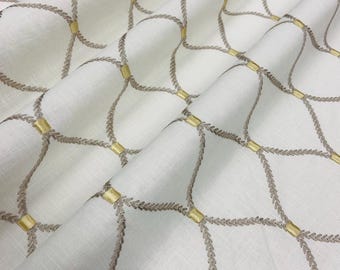 Può includere: Tessuto bianco con un motivo ripetuto di linee ricamate a forma di corda di colore marrone chiaro, collegate da piccoli quadrati dorati. Il tessuto è piegato, mostrando la trama e il design.