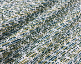 Puede incluir: Tejido textil con un patrón tejido de guiones azules, verdes y dorados sobre un fondo blanco. La tela tiene una apariencia texturizada, con un ligero brillo. El diseño se repite en la superficie, creando un efecto geométrico.