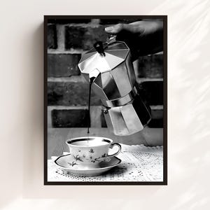 May include: Black and white photograph of coffee being poured from a silver stovetop espresso maker into a delicate teacup and saucer. The cup and saucer have a floral design. The background features a brick wall.