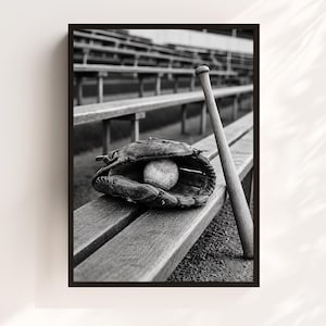 Op de afbeelding: Een zwart-wit ingelijste wandkunstprint met een leren honkbalhandschoen die een honkbal vasthoudt, rustend op een houten stadionbank naast een honkbalknuppel, met wazige stadiontribunes zichtbaar op de achtergrond.