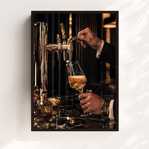 May include: A framed image of a person pouring a drink from a gold tap into a wine glass. The glass is held by a person wearing a black suit and a watch. The background is dark with gold accents.