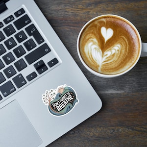 Może przedstawiać: Laptop z naklejką z napisem "Don't Forget to Breathe" obok filiżanki kawy z latte art. Naklejka przedstawia słońce, fale i drzewo. Kawa ma wzór w kształcie serca.