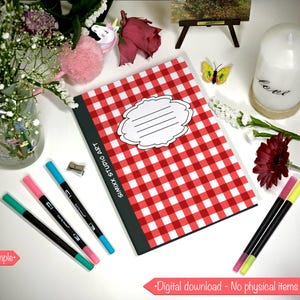 May include: A red and white checkered notebook with a blank label is the focal point of this image. Several colorful markers, a candle, and flowers are arranged around the notebook. The text "SIMIXX STUDIO ART" is printed on the notebook.