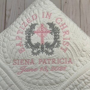 May include: White quilted baby blanket with pink and gray embroidery. The embroidery reads "Baptized in Christ" with a cross and floral design. The name "Siena Patricia" and the date "June 13, 2021" are also embroidered on the blanket.