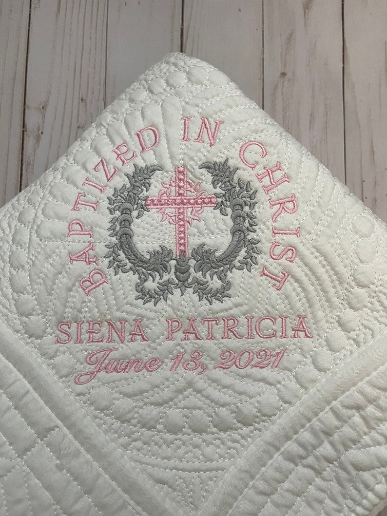 May include: White quilted baby blanket with pink and gray embroidery. The embroidery reads "Baptized in Christ" with a cross and floral design. The name "Siena Patricia" and the date "June 13, 2021" are also embroidered on the blanket.
