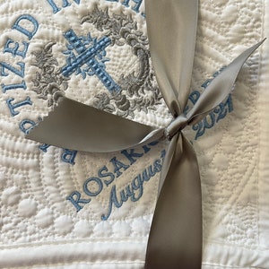 May include: White quilted baby blanket with a gray satin ribbon tied in a bow. The blanket is embroidered with a cross, a wreath, and the text "Baptized in Christ" and "Rosakko August 2021".