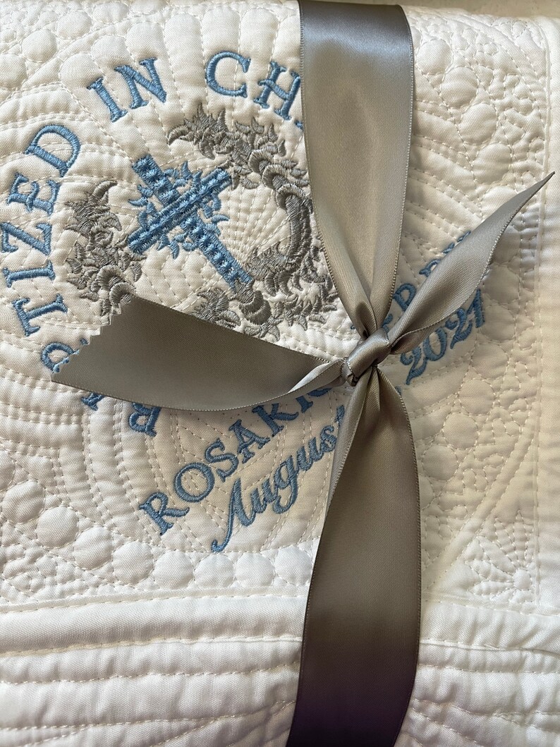 May include: White quilted baby blanket with a gray satin ribbon tied in a bow. The blanket is embroidered with a cross, a wreath, and the text "Baptized in Christ" and "Rosakko August 2021".