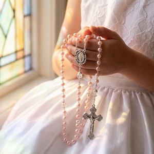 May include: A pink rosary with pearl-like beads, a silver crucifix, and a decorative centerpiece. The rosary is held in front of a white dress near a stained-glass window.