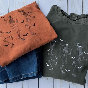 Pode incluir: Duas sweatshirts com tema de Halloween. Uma &eacute; laranja com esqueletos e morcegos pretos, a outra &eacute; verde escura com esqueletos e morcegos brancos.