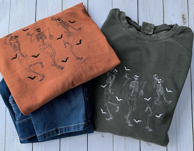 Pode incluir: Duas sweatshirts com tema de Halloween. Uma &eacute; laranja com esqueletos e morcegos pretos, a outra &eacute; verde escura com esqueletos e morcegos brancos.
