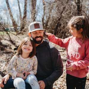 Peut inclure: Un homme portant une casquette de baseball grise avec le texte "DAD LIFE" en lettres brunes est assis avec deux jeunes filles. La fille sur les genoux de l'homme porte une chemise &agrave; fleurs jaunes et la fille debout porte un sweat-shirt rose avec le texte "PINK FLOYD" en lettres blanches.