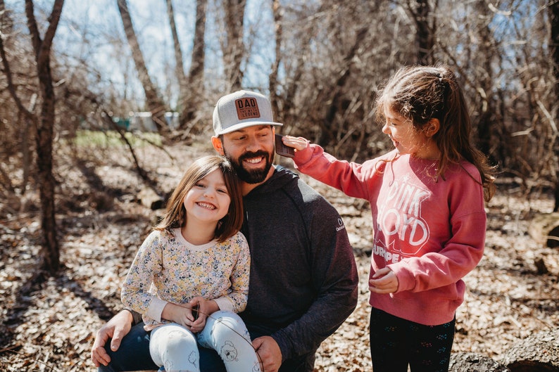Peut inclure: Un homme portant une casquette de baseball grise avec le texte "DAD LIFE" en lettres brunes est assis avec deux jeunes filles. La fille sur les genoux de l'homme porte une chemise &agrave; fleurs jaunes et la fille debout porte un sweat-shirt rose avec le texte "PINK FLOYD" en lettres blanches.