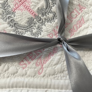 May include: A white quilted baby blanket with a silver ribbon tied in a bow. The blanket is embroidered with a pink cross, a floral design, and the text "SIENA" and "June" in pink thread. The text "PATRICIA 2021" is also embroidered in pink thread.
