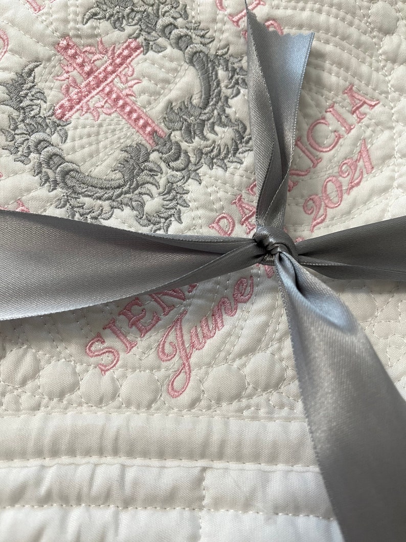 May include: A white quilted baby blanket with a silver ribbon tied in a bow. The blanket is embroidered with a pink cross, a floral design, and the text "SIENA" and "June" in pink thread. The text "PATRICIA 2021" is also embroidered in pink thread.