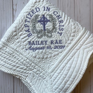 May include: White quilted baby blanket with embroidered text "Baptized in Christ" and a cross design. The text "Bailey Rae August 15, 2021" is also embroidered on the blanket.