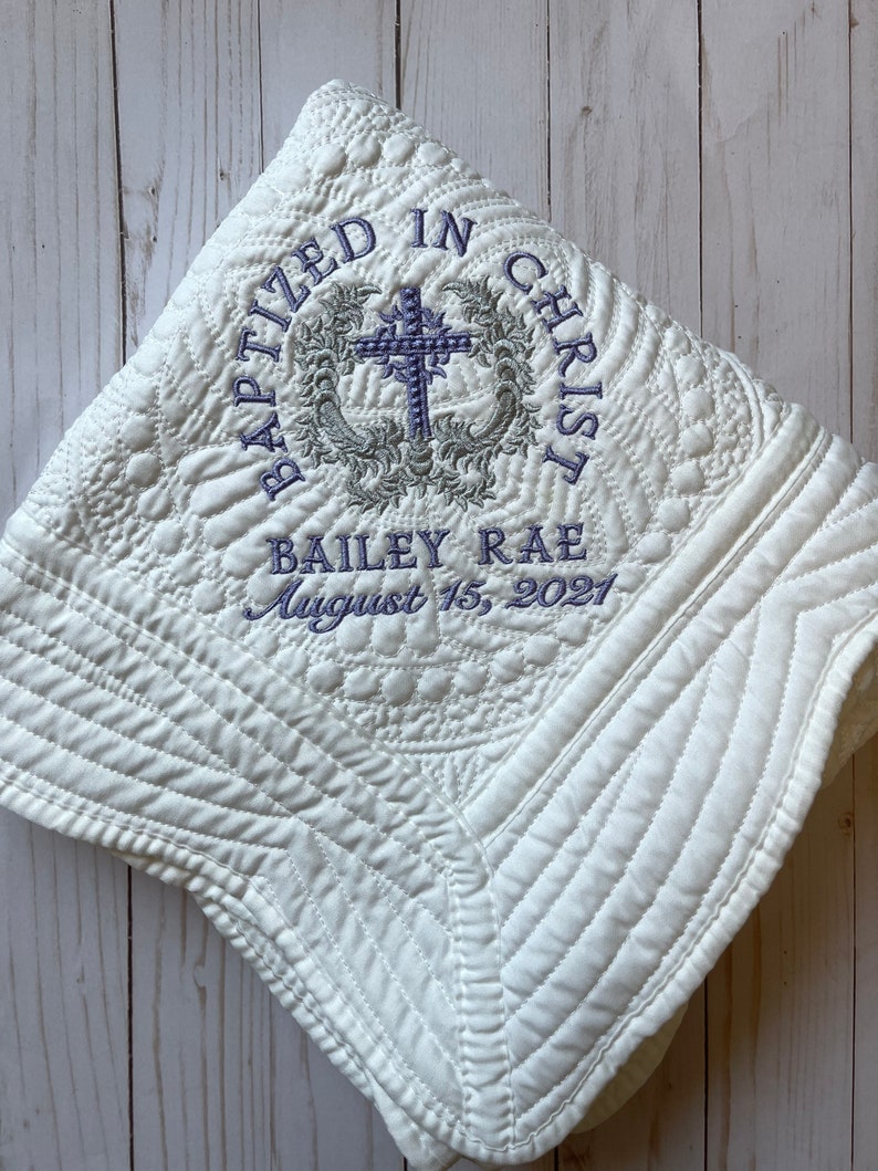May include: White quilted baby blanket with embroidered text "Baptized in Christ" and a cross design. The text "Bailey Rae August 15, 2021" is also embroidered on the blanket.