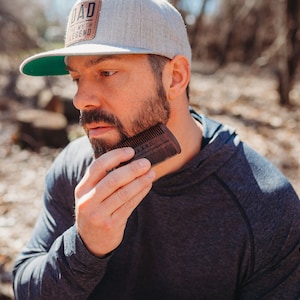 Peut inclure: Un homme barbu se coiffe la barbe avec un peigne &agrave; barbe en bois. Le peigne est grav&eacute; des mots "Bearded" et "The Art of Grooming Your Beard".