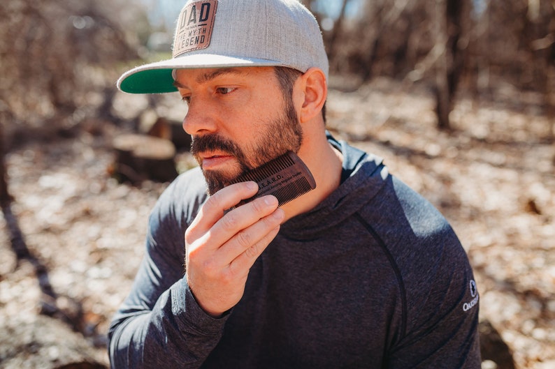 Peut inclure: Un homme barbu se coiffe la barbe avec un peigne &agrave; barbe en bois. Le peigne est grav&eacute; des mots "Bearded" et "The Art of Grooming Your Beard".